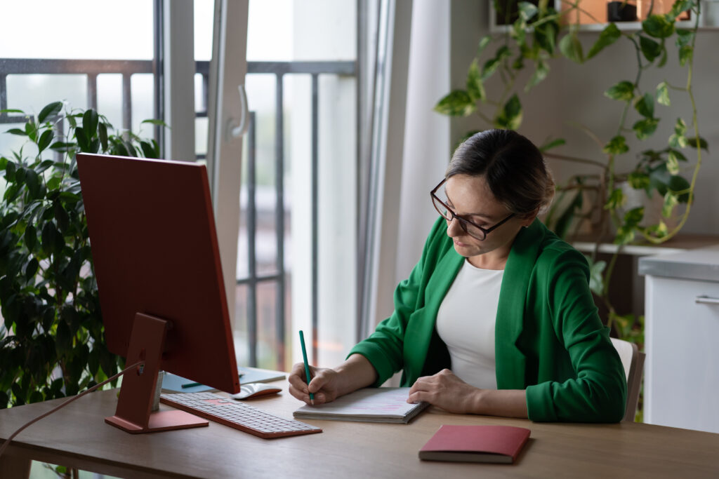 Focused Caucasian woman tutor or teacher making notes in notebook sits at table with computer